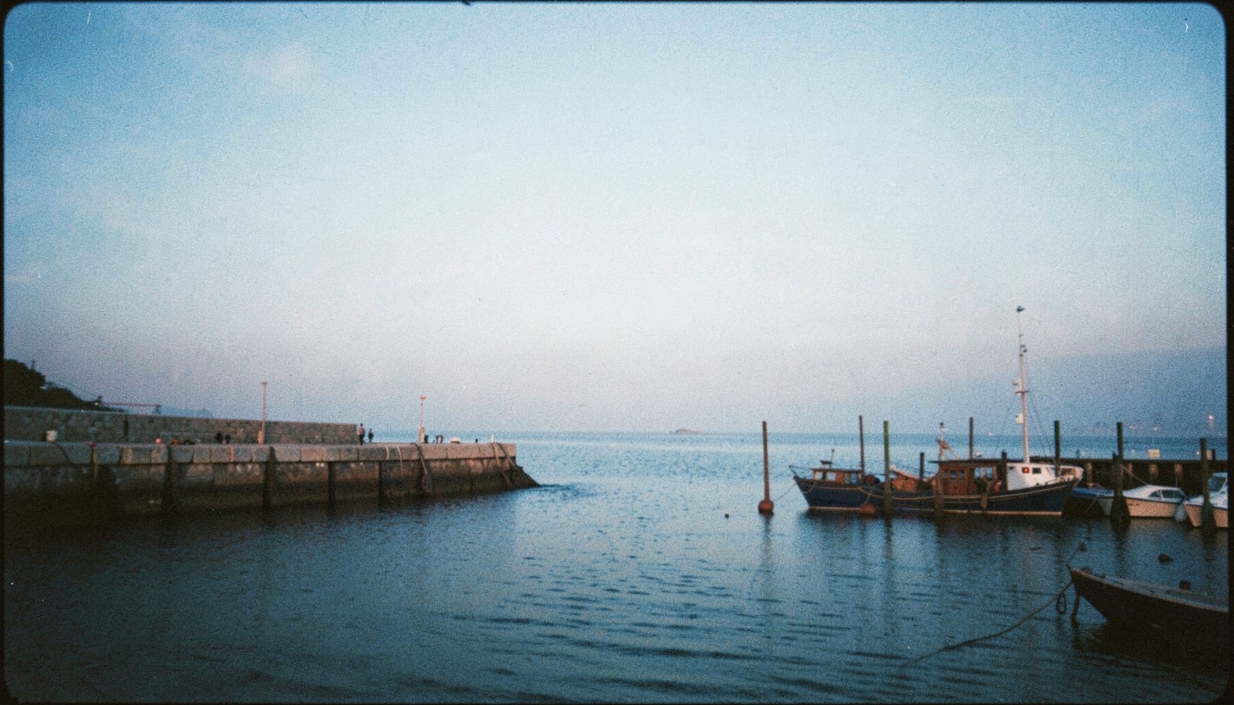 Wide analog film photograph of a quiet coastal harbor at dusk, with soft grain, pale blue sky fading into gray water, a low stone breakwater, and a few moored wooden boats in muted brown and rust tones